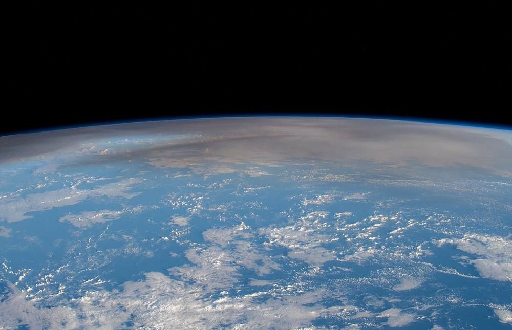 Earth's curved horizon seen from orbit, with a thin high-altitude plume layer above the clouds.