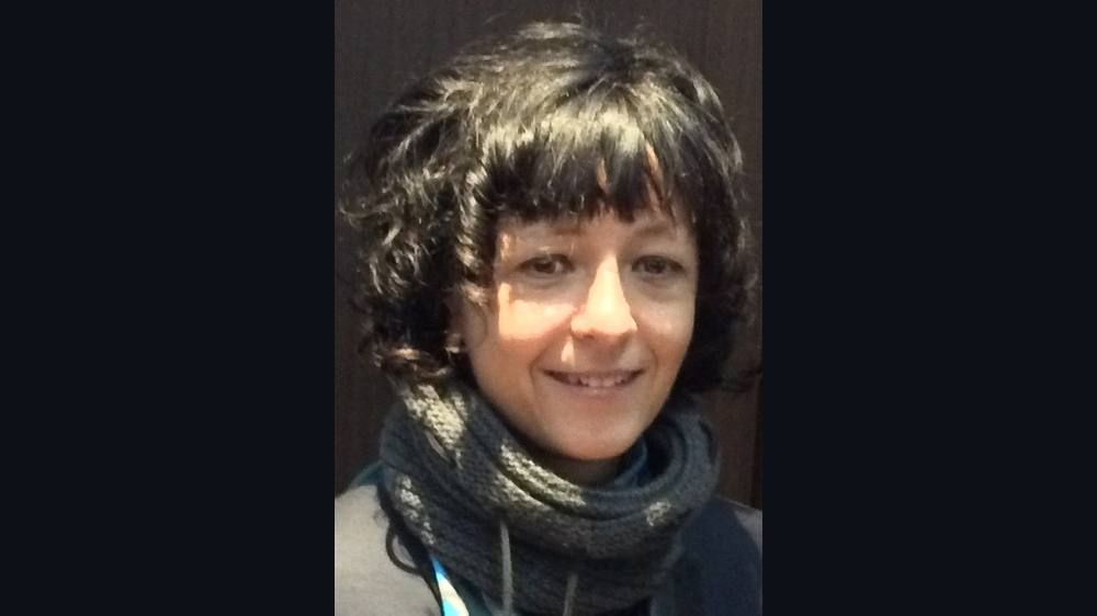 A smiling woman with dark curly hair and a scarf, photographed outdoors.