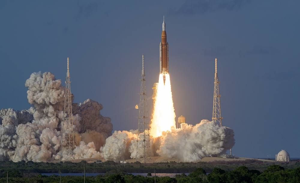 A tall orange and white rocket rises on a blaze of flame and clouds of smoke from its launch pad.