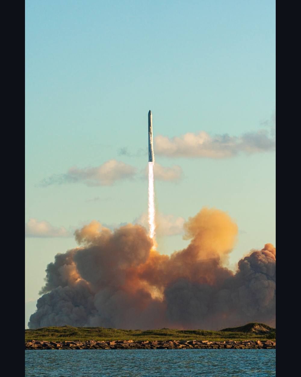 A Starship stack climbs into a blue sky on a column of fire above its launch site.