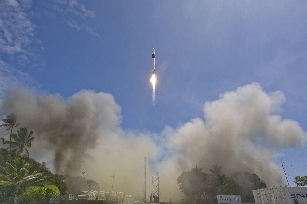 A small white rocket climbs from a tropical island launch site with a bright exhaust flame and a plume of smoke.