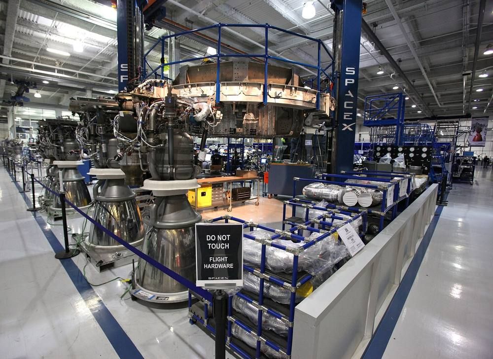 Several rocket engines with silver bells stand on the floor of a factory next to a large metal engine mounting frame.