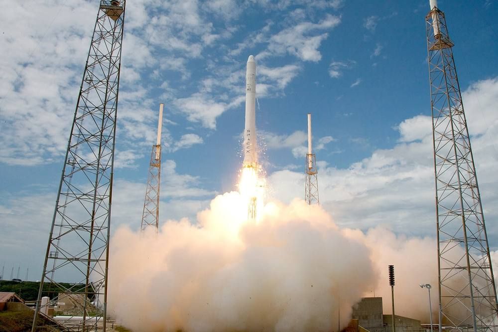 A white rocket rises from its pad with a bright exhaust flame and a trail of smoke against a pale sky.