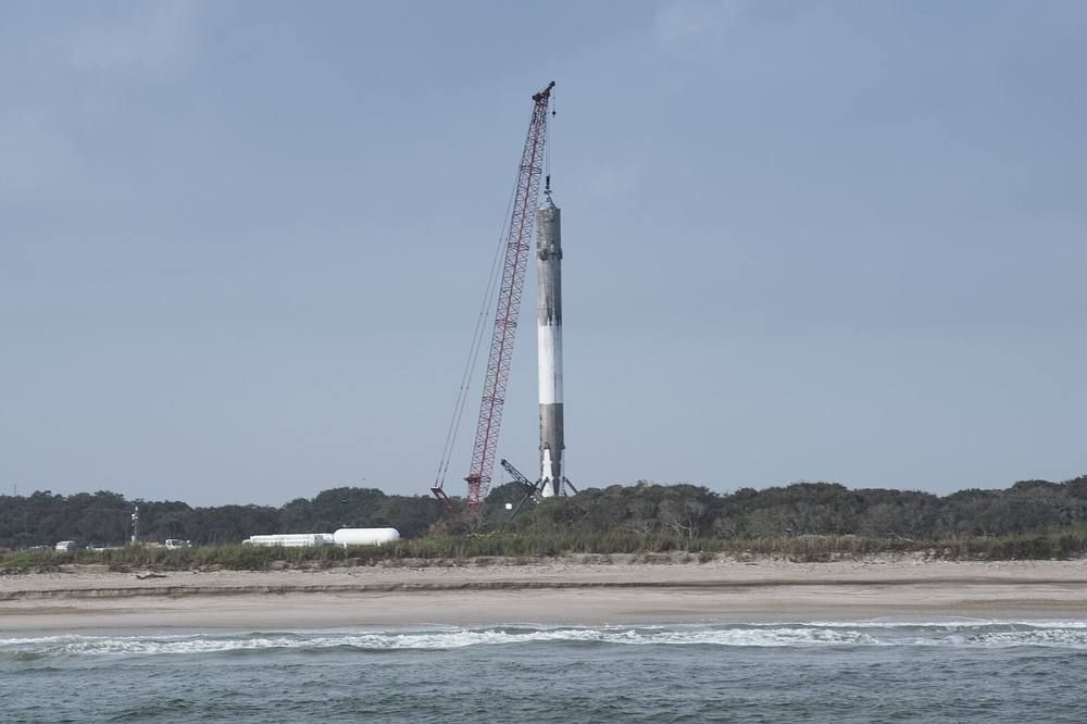 A tall white booster with landing legs stands upright on a concrete pad by the coast under a pale sky.