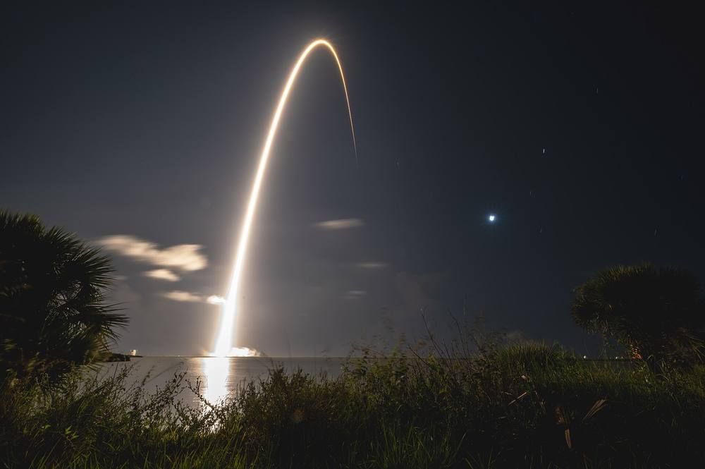 A long bright arc of light rises over dark trees and water in a night time-exposure of a rocket launch.