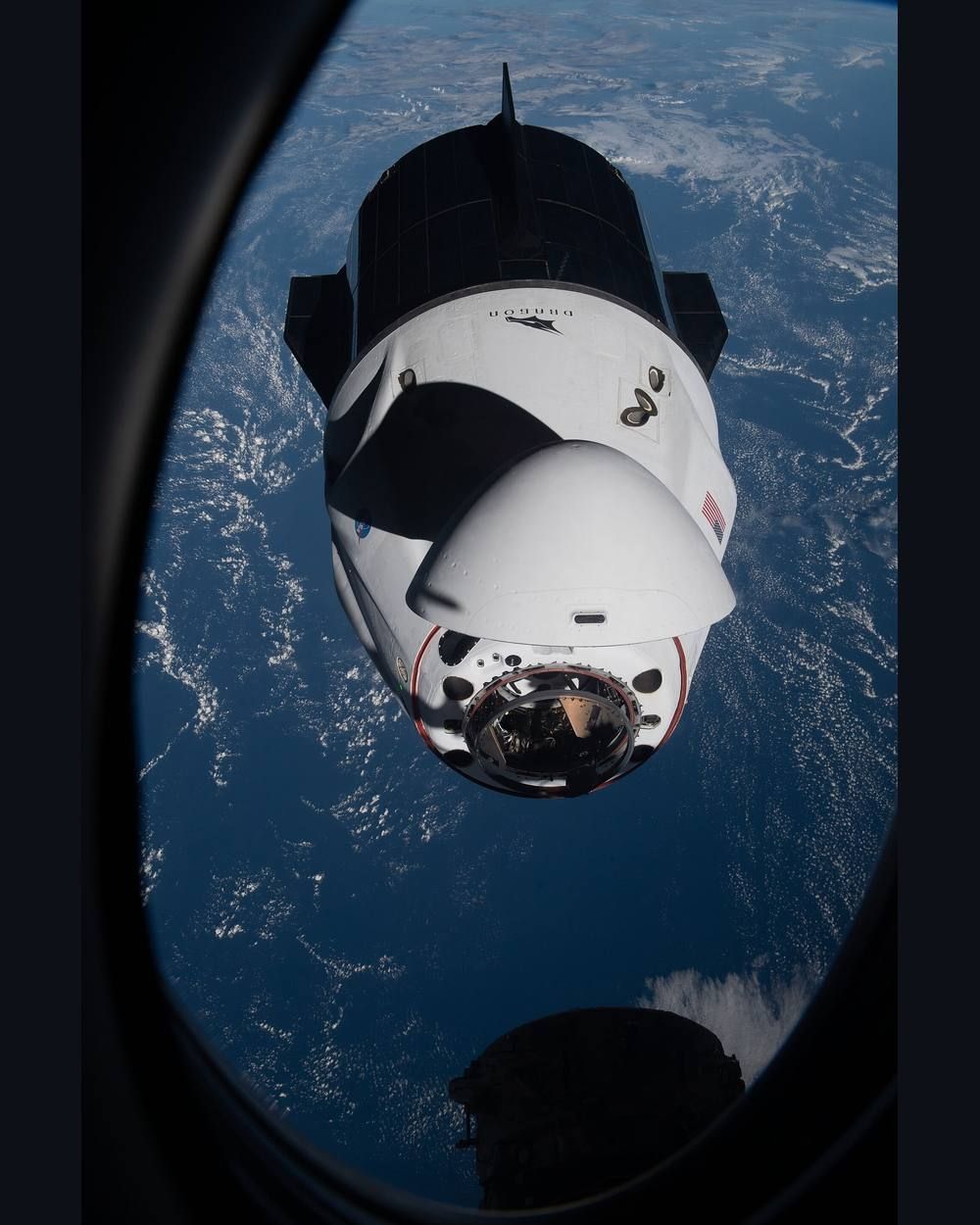 A white and black spacecraft with solar-paneled trunk and a docking nose floats against the black of space with Earth below.