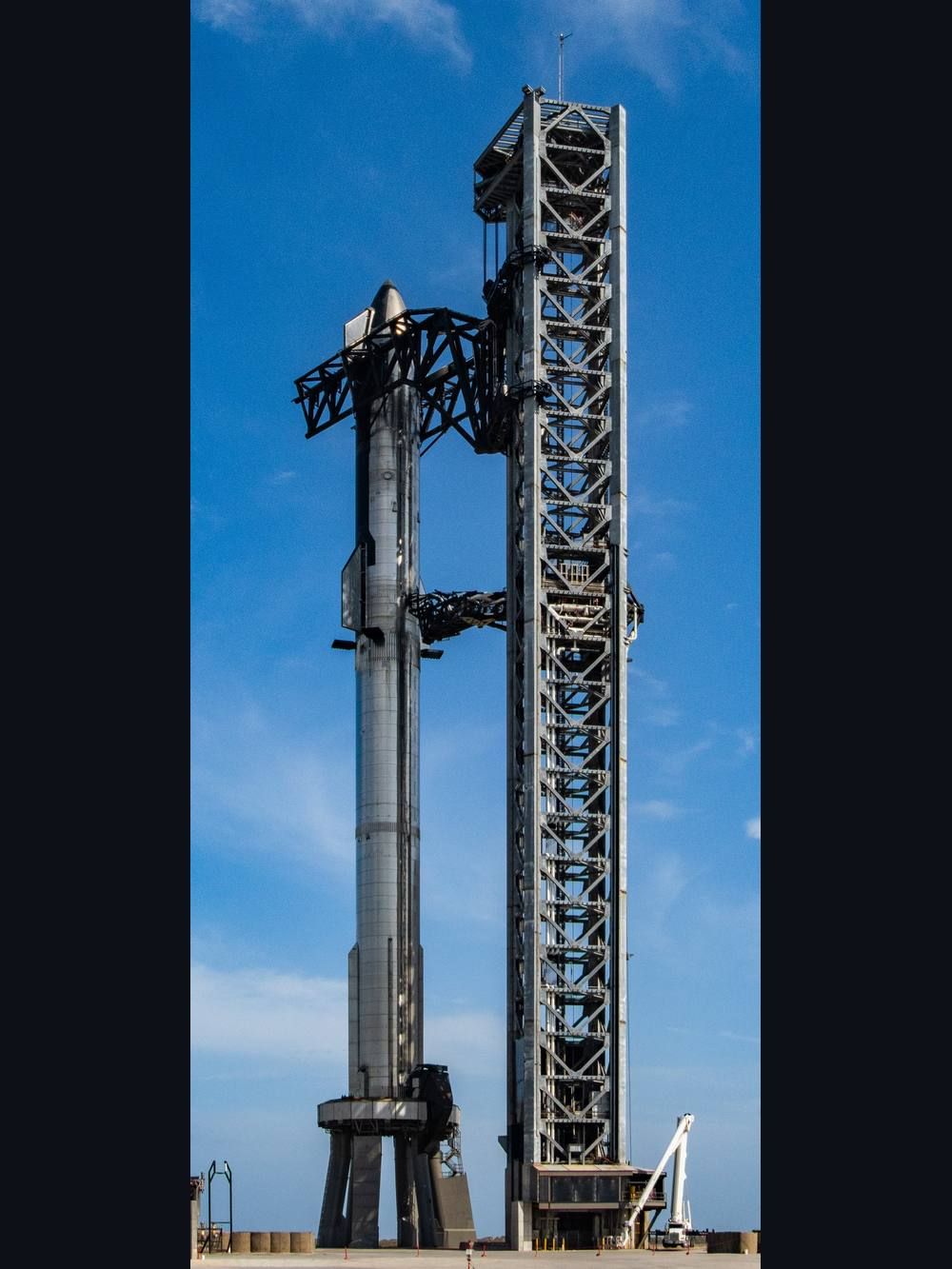 A tall stainless-steel rocket stack stands beside a lattice launch tower against a blue sky.