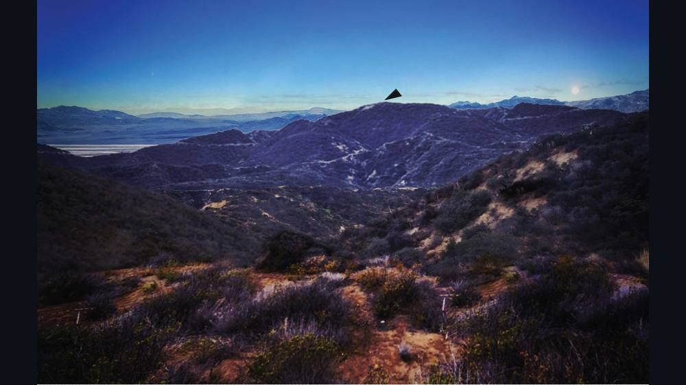 A dusk landscape of dry brush and ridges with a mountain skyline and a small flat black triangle shape above the central ridge.